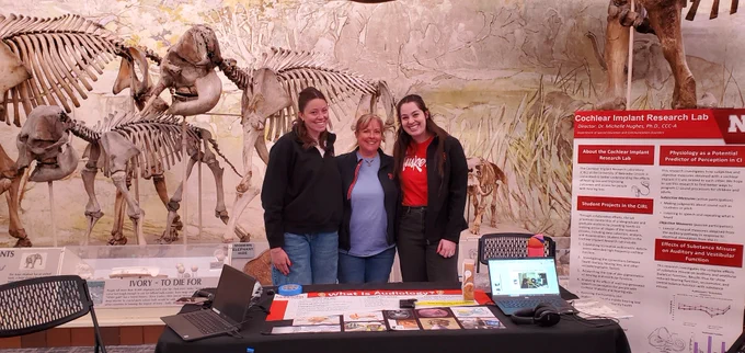Michelle Hughes poses with two student researchers behind a table for the Cochlear Implant Research Lab with ancient elephant skeletons n the background