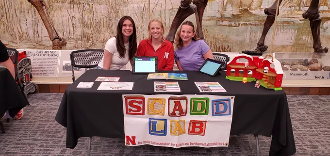 three student researchers pose for a photo together behind a table with a black tablecloth and a white sign on front with SCADD LAB in spelled out in different colored children's blocks