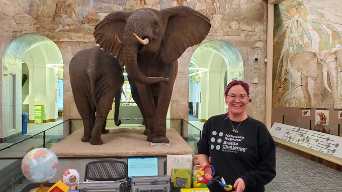 Mackenzie Savaiano holds a device in her hands next to a table of equipment and toys related to teaching students with visual impairments while wearing a black sweatshirt that says "Nebraska Regional Braille Challenge" in front of two elephant sculptures in Morrill Hall