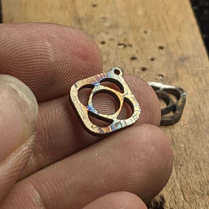 Holding a tiny rounded-square pendant (the Psyche mission logo) cut from meteorite. A second piece lays on the wooden bench behind it.