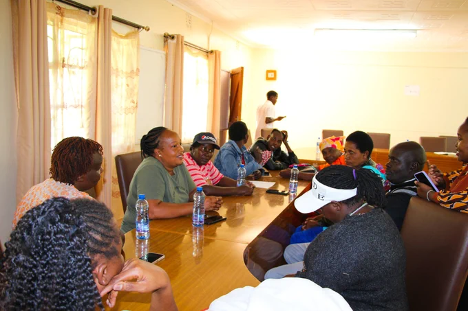 The image shows a community engagement session taking place indoors. A group of parents is seated around a large table, actively participating in a discussion. One woman appears to be facilitating the session, speaking while others listen attentively, some leaning in.