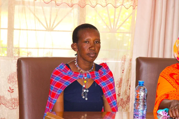 This image shows a focused moment within a community dialogue session. The woman seated at the table, attentive and reflective, listening to the discussion. Her traditional attire and beaded accessories suggest cultural identity and community representation.

