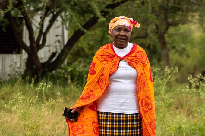  A woman stands outdoors, surrounded by greenery, holding a phone and dressed in vibrant traditional attire, including a patterned wrap and headscarf. Her posture and expression suggest confidence, dignity, and a sense of purpose.