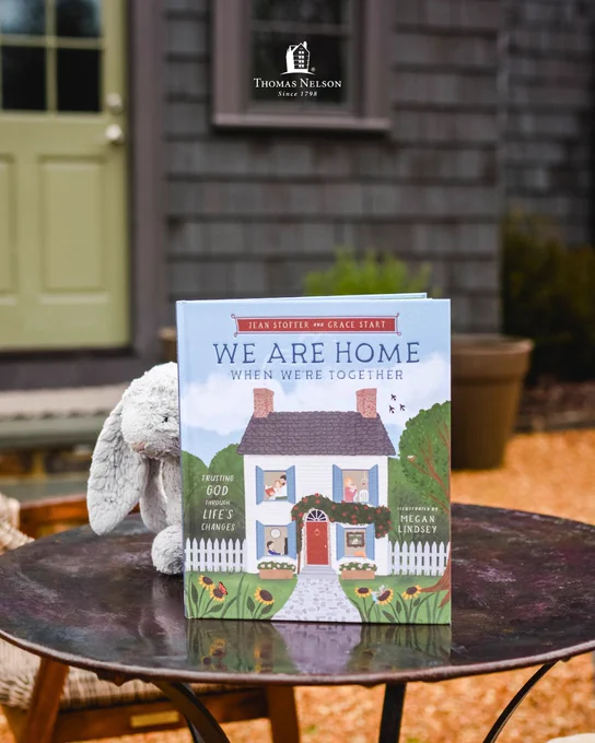 Book titled "We Are Home: When We're Together" by Jean Stoffer, Grace Stoffer, and Megan Lindsey, on a round outdoor table. The cover depicts a charming house with a pathway, surrounded by greenery. A plush gray rabbit is partially visible beside the book. Thomas Nelson logo is visible above.
