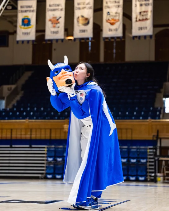 Duke student Nana Osaki posing in Cameron Indoor Stadium. She is wearing the body of the Blue Devil mascot costume, and holding the Blue Devil mascot head, giving it a kiss on the cheek. Behind Nana, you can see Duke Men's Basketball team's five National Champions banners hanging from the rafters. 