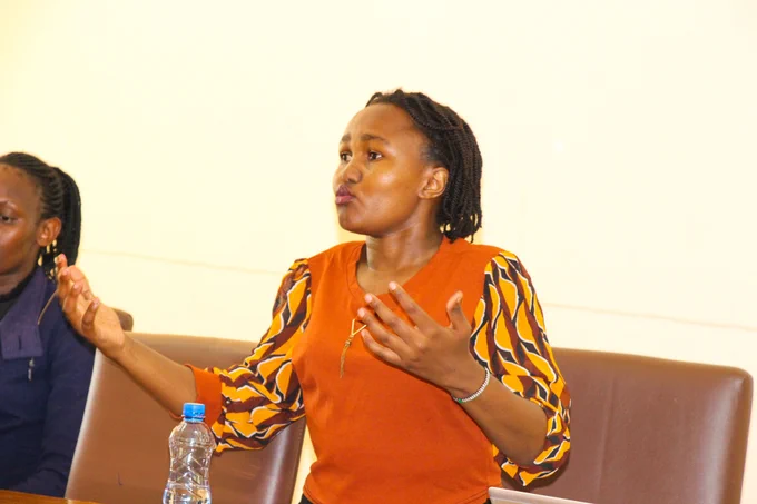 In the foreground, a young Black woman with braided hair is standing, captured in a mid-motion pose as she speaks. She is wearing an orange, V-neck shirt with voluminous sleeves that feature a striking, abstract pattern of yellow, brown, and black shape.Her right hand is raised and open, with fingers spread. Her left hand is also slightly raised, with fingers tucked in. Her facial expression is animated, with pursed lips and focused eyes looking to her left, indicating she is actively engaged in conversation. 