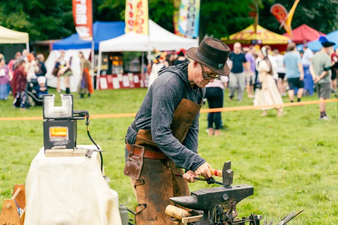A person dressed in a hat, glasses, and a leather apron is engaged in metalworking at an outdoor fair with colorful tents and a crowd of people visible in the background on a grassy field.