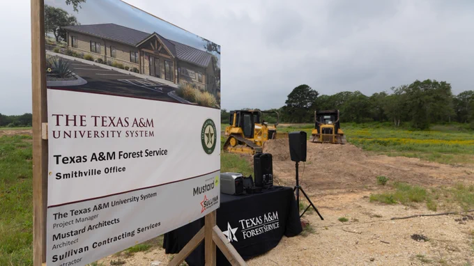 A construction site with a Texas A&M Forest Service sign indicating the location of a future Smithville Office. The sign mentions project partners: Mustard Architects and Sullivan Contracting Services.