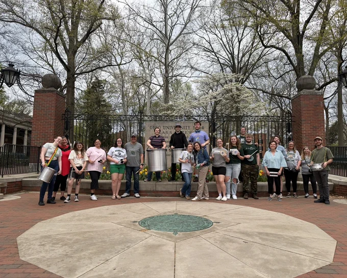 A group of people standing in front of an Ohio University gateway, surrounded by trees. Some are holding drums. It appears to be an outdoor gathering, with a brick pathway in the foreground.