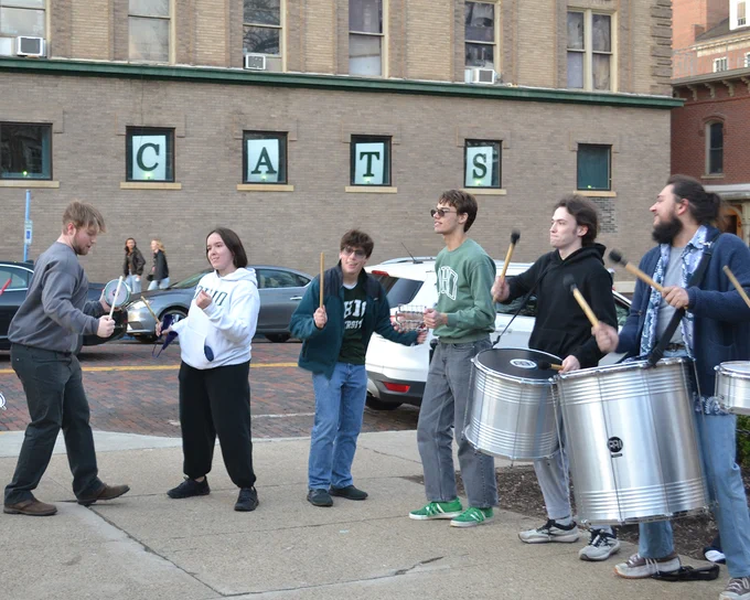 A group of people is playing percussion instruments on a sidewalk. They are standing in front of a building with large letters spelling "CATS" in the windows. The scene conveys a lively outdoor performance.