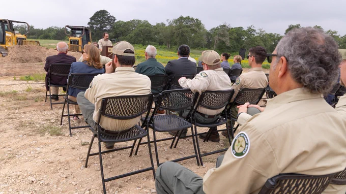 A group of people are seated outdoors on folding chairs, listening to a speaker. They are near construction equipment, including bulldozers, in a natural setting with trees in the background. Most attendees are wearing uniforms with patches, and one is speaking at the front.