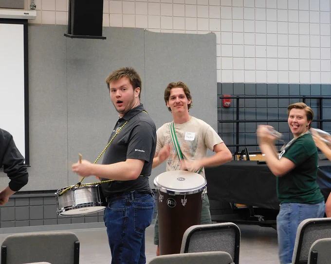 Three people in a classroom setting are playing percussion instruments. The person in the middle plays a drum, while the others shake handheld instruments. They seem engaged and enthusiastic.