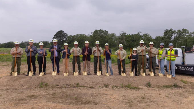A group of individuals wearing helmets and holding shovels stand in front of a mound of dirt at a groundbreaking ceremony. The people are dressed in a mix of formal and work attire. In the background, there is a vehicle with the Texas A&M Forest Service logo. 