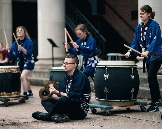 A group of individuals is performing a traditional drum ensemble in front of a building. They are wearing blue and white garments and playing large taiko drums and small cymbals. The scene conveys energy and coordination.