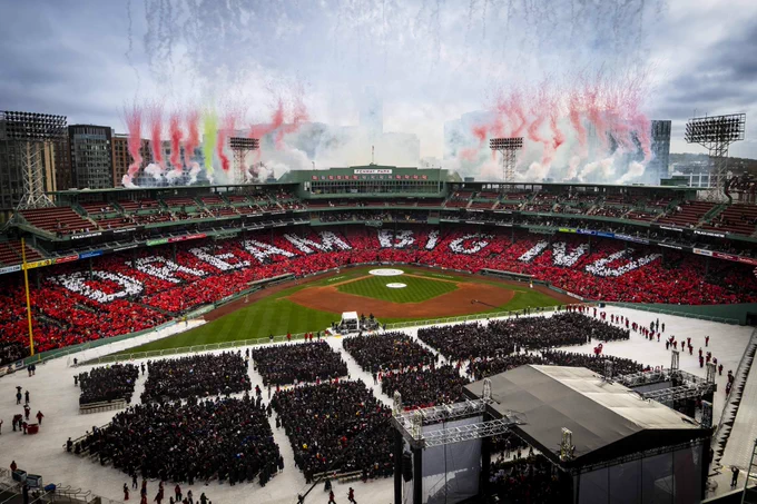 Northeastern University 2026 commencement at Fenway Park with \"DREAM BIG\" formed by fans in red and white. Graduates sit in black robes. Above, celebratory red and green smoke rises.