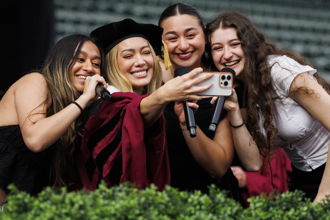 Undergraduate Commencement speaker Hilary Duff poses for a selfie with three students, who are singing on stage. Duff wears graduation attire, including a black cap with gold tassel and a maroon gown. The students hold microphones. 