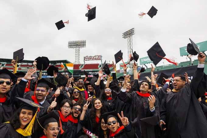 Graduates in black gowns joyfully toss caps at Fenway Park stadium, celebrating with raised hands and peace signs. 