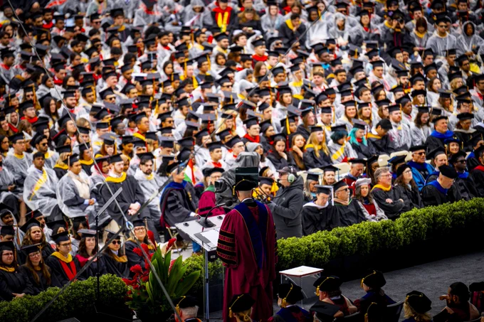 A speaker in a maroon robe stands at a podium addressing a large crowd of graduates in caps and gowns. Some attendees are wearing gray ponchos. Red flowers adorn the stage.