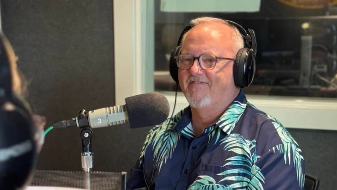 Dr. Roger Smith with headphones sits in a radio studio, speaking into a microphone. They are wearing a blue shirt with a palm leaf pattern.