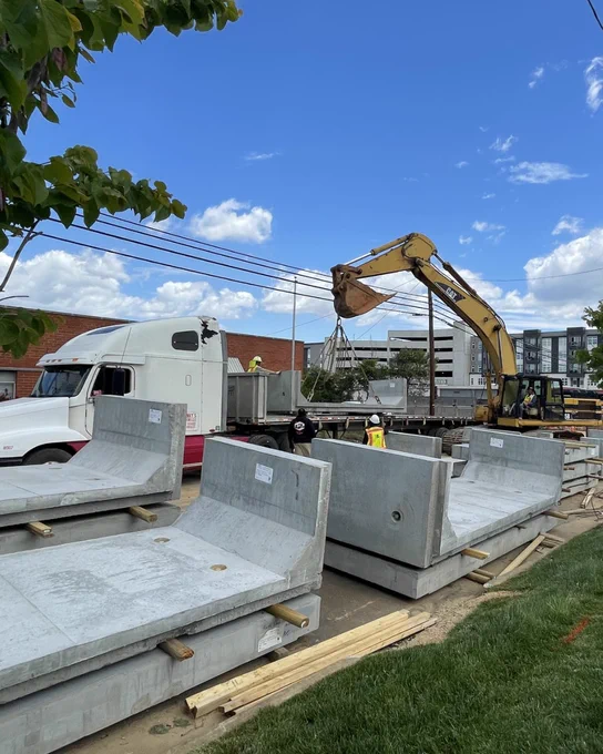 A construction site with a large excavator moving concrete barriers. Workers are guiding the equipment near a parked semi-truck. In the background are buildings and a partly cloudy blue sky.