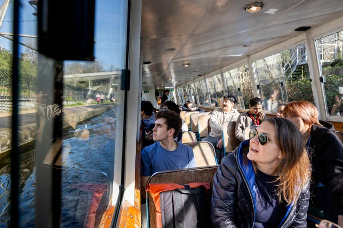 A canal boat interior view shows a group of students in natural light. A woman in sunglasses and a blue jacket is in focus. Reflections on the window show the canal and onlookers.