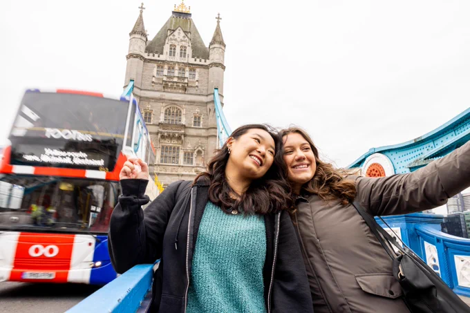 Two smiling women pose on a bridge with Tower Bridge in the background. A red double-decker bus passes them,  blurred in motion. The women \u2013 both students \u2013  are dressed in casual jackets and sweaters.