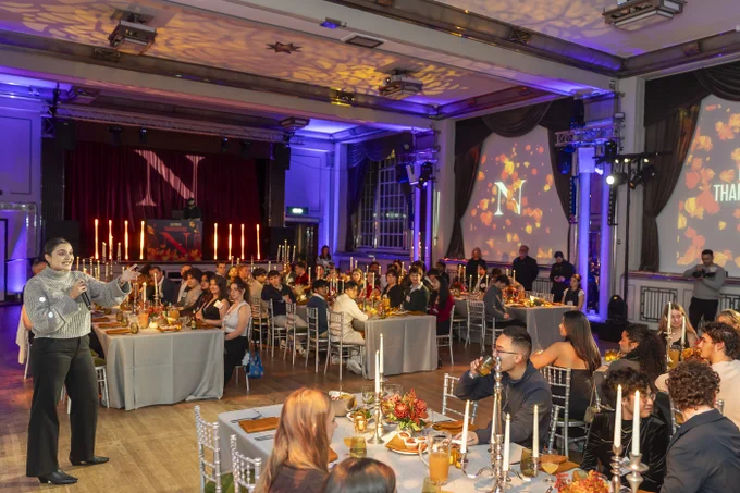In a banquet hall, a female student speaks into a microphone before seated student guests at tables set for a meal. A stage with a screen displaying \"N\" and \"THANKSGIVING\" is behind the audience. Candles and floral arrangements decorate the tables.