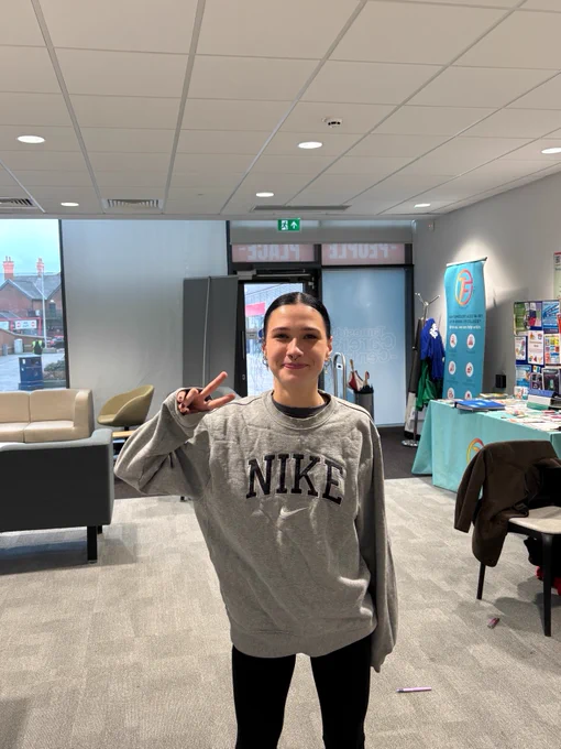 A person stands indoors, wearing a grey Nike sweatshirt and making a peace sign. 