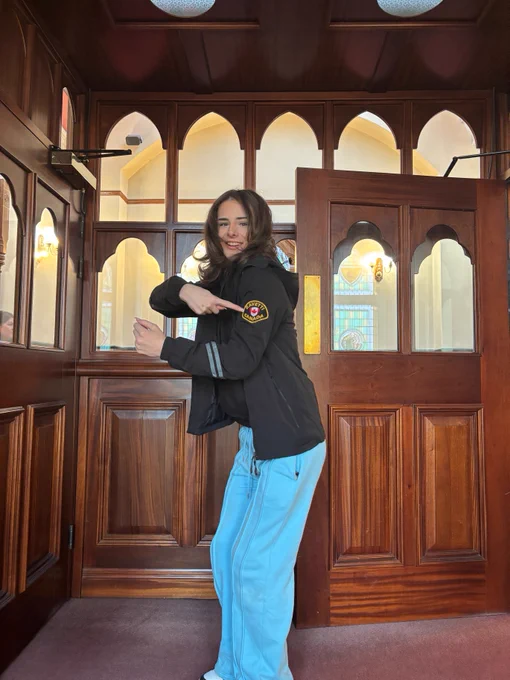A person in a black jacket and light blue pants stands in a wooden room with arched windows and doors, pointing at a patch on the jacket sleeve.
