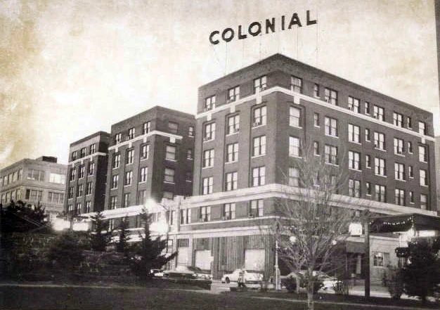 Historic photograph of the Colonial Hotel, a multi-story brick building with large windows. The name "COLONIAL" is prominently displayed on the roof. There are trees and parked cars in the foreground.