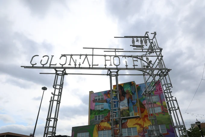 A large metal sign reads "Colonial Hotel" against a cloudy sky. Below the sign, a colorful mural decorates a multi-story building.