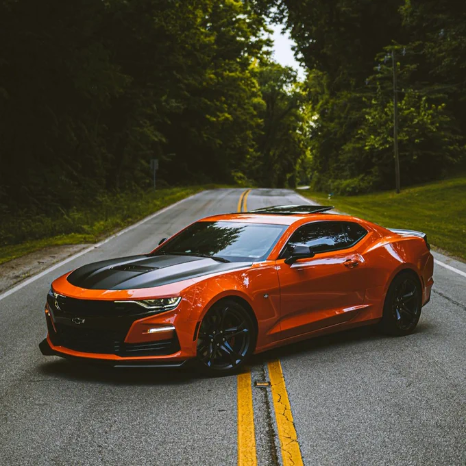 An orange sports car with a black hood parked on a tree-lined road with double yellow lines.