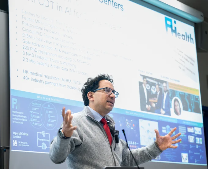 Professor Aldo Faisal speaks at a lectern during a presentation on AI for healthcare. He is standing in front of a large projected slide, with his hands raised as he addresses the audience. The slide includes information about AI and healthcare centres, doctoral training, NHS hospital trusts, patient data, industry partners and the UKRI Centre for Doctoral Training in Artificial Intelligence for Healthcare at Imperial College London.