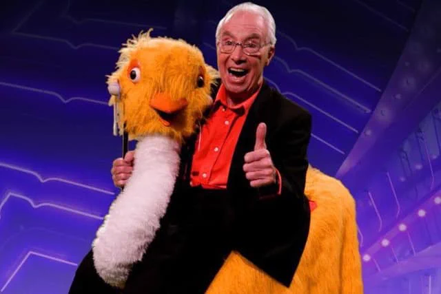 Bernie Clifton wearing suit and glasses, giving a thumbs and smiling whilst astride his trusty ostrich 