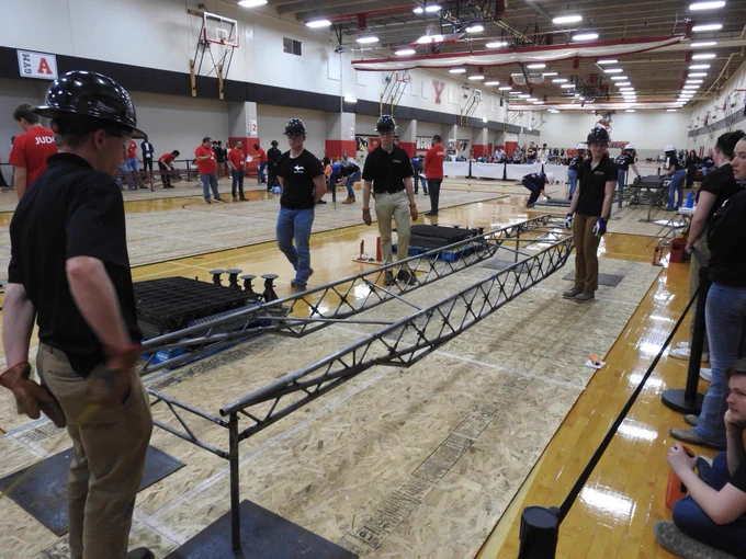 Michigan Tech students wearing helmets assemble a long metal truss structure on a gymnasium floor during a team event.