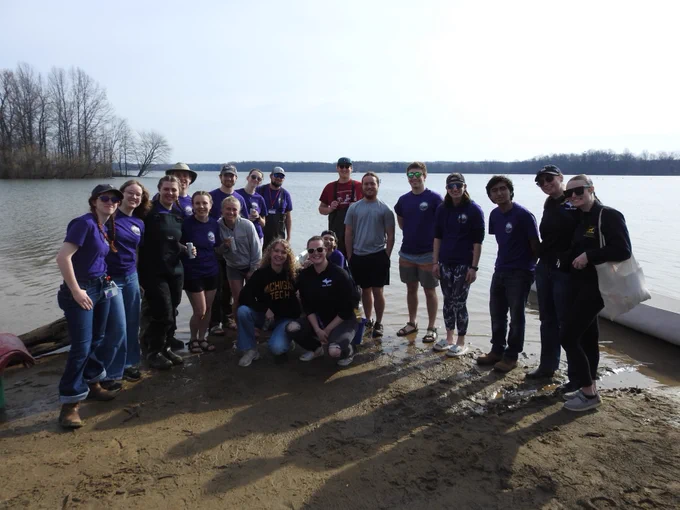 Group of Michigan Tech students standing on a sandy shore by a calm lake on a clear day, some wearing purple shirts and casual outdoor clothing.