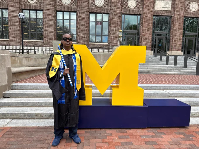 Graduate student in full regalia standing in front of a giant block M in front of the U-M Hatcher Library.