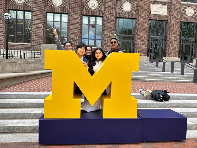 Happy family standing behind a giant block M in front of the U-M Hatcher Library.