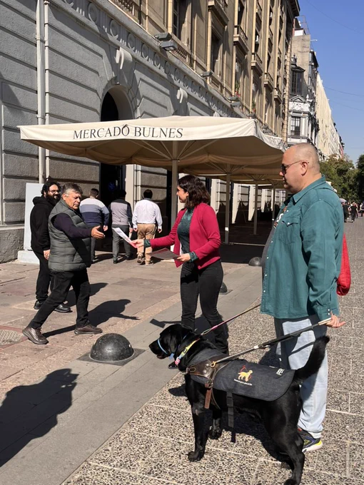 Fotografía en la que aparece la Seremi de Desarrollo Social y Familia, junto a Mauricio Flores y su perro guía choco, entregando información a la ciudadanía con recomendaciones para el respeto de personas usuarias de perro guía.