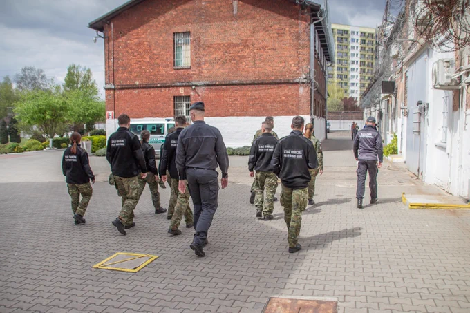 Umundurowani funkcjonariusze Straży Granicznej spacerują po terenie ścisłym jednostki penitencjarnej w asyście umundurowanych funkcjonariuszy Służby Więziennej.