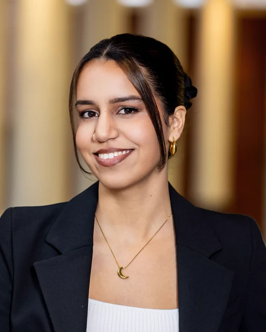 woman with dark hair pulled back, smiling, wearing a black blazer and white shirt with gold necklace and earrings.