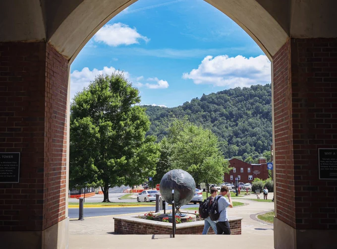 A sunny campus scene that is framed by “brick legs” of a bell tower. Students are walking by in front of a globe sculpture surrounded by flowers. Lush, green mountains are visible in the distance.