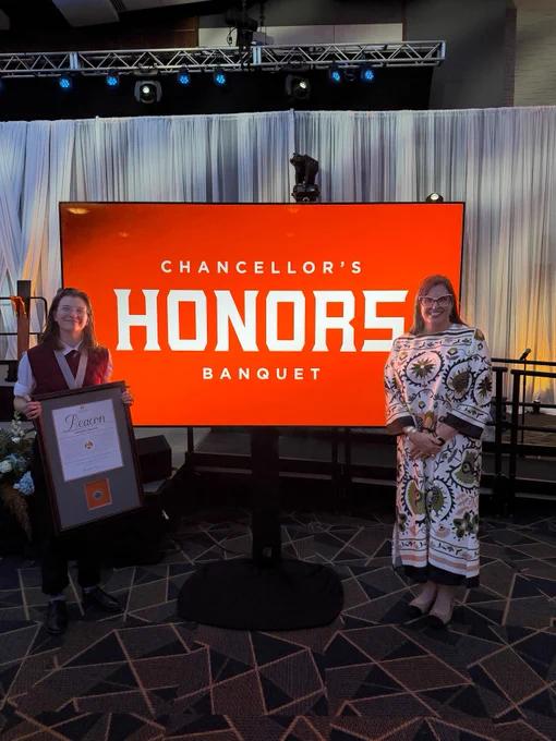 Beacon award winner Lauren Bailey standing on one side of an orange sign with the words "Chancellor's Honors Banquet" with Marieke Van Puymbroeck, Dean of the Graduate School, on the opposite side.