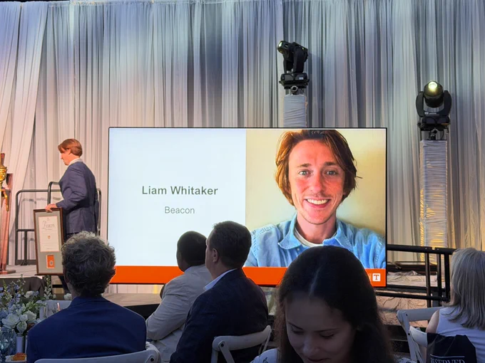 Liam Whitaker taking the stage with his Beacon award, near a large display with a photo of him next to his name.