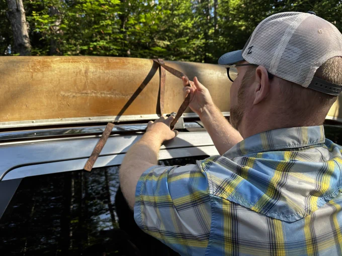 A person wearing a cap and glasses secures a weathered canoe onto the roof of a vehicle using straps.