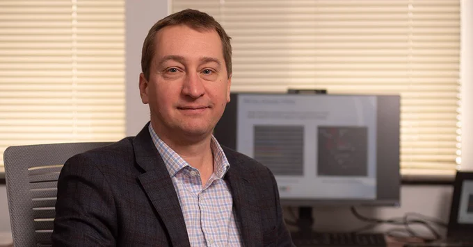 Professional headshot of Chris Hinkle seated at a desk in an office, with a computer monitor showing research visuals in the background.