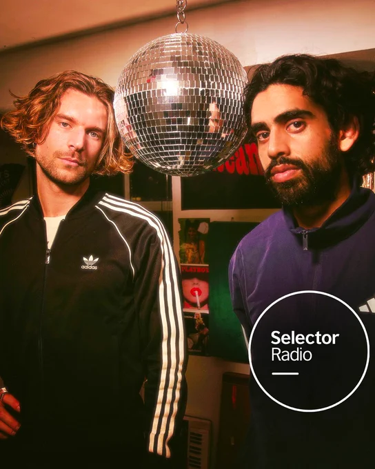 Music duo KuleeAngee (Duncan Grant and Keshav Kanabar) stand close together indoors, framed by a disco ball overhead and a wall of posters behind them. Their expressions are calm and focused, dressed in sporty jackets that reflect a casual style. The lighting gives the image a vintage, intimate studio vibe as they look into the camera. The Selector Radio logo appears in a white circle in the bottom right corner.