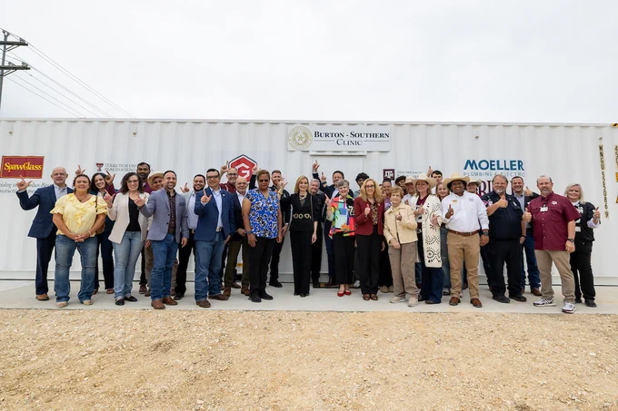 A group of over 20 professionals stands in front of a white shipping container with a sign that says "Burton‑Southern Clinic." The group is posing for a photo outside under a cloudy sky. Some individuals are giving thumbs up, or "Gig 'em" signs, while others are giving the Red Raiders sign.