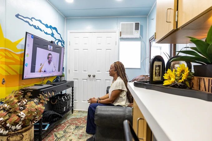 A person sits on a chair in a colorful room, engaged in a video call displayed on a large television screen. The room features vibrant wall art and plants. A shelf with decorative items is visible on the right.