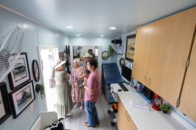 A group of people in a brightly lit mobile health clinic with light blue walls and wooden cabinets. Some historic photographs and health-related posters are on the walls. A bench, medical examination table, and various medical equipment are visible.
